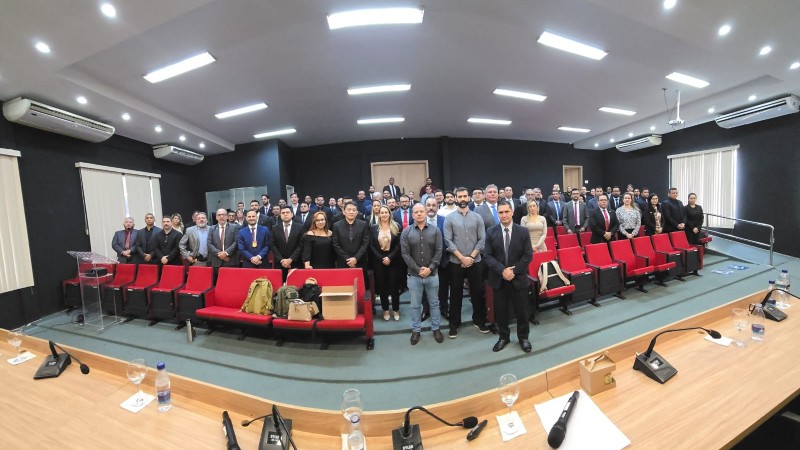Audit&oacute;rio, com autoridades e convidados em trajes sociais posicionados atr&aacute;s de uma mesa equipada com microfones e copos de &aacute;gua, enquanto o p&uacute;blico ocupa cadeiras vermelhas, refor&ccedil;ando o car&aacute;ter institucional da ocasi&atilde;o.
