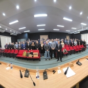 Audit&oacute;rio, com autoridades e convidados em trajes sociais posicionados atr&aacute;s de uma mesa equipada com microfones e copos de &aacute;gua, enquanto o p&uacute;blico ocupa cadeiras vermelhas, refor&ccedil;ando o car&aacute;ter institucional da ocasi&atilde;o.