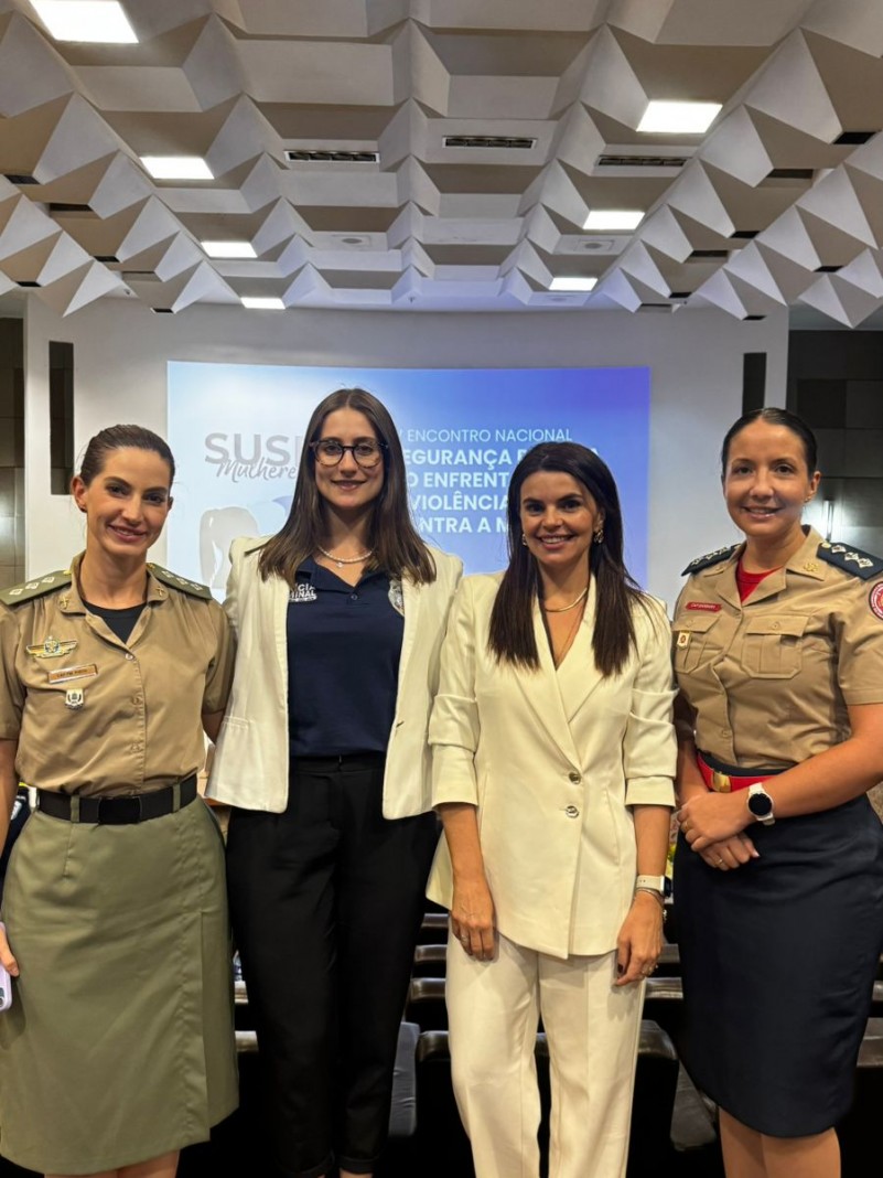 Quatro mulheres est&atilde;o em p&eacute;, lado a lado, em uma sala de confer&ecirc;ncia ou semin&aacute;rio. Duas delas vestem uniformes de for&ccedil;as de seguran&ccedil;a (militares ou policiais), enquanto as outras duas usam roupas profissionais civis. Ao fundo, h&aacute; uma tela de proje&ccedil;&atilde;o exibindo um slide com o texto em portugu&ecirc;s:
"SUS Mulheres &ndash; 1&ordm; Encontro Nacional &ndash; Seguran&ccedil;a P&uacute;blica e o Enfrentamento da Viol&ecirc;ncia Contra a Mulher". O ambiente transmite formalidade e colabora&ccedil;&atilde;o entre representantes da seguran&ccedil;a p&uacute;blica e da sociedade civil.