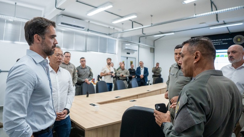 Amplo sagu&atilde;o com v&aacute;rias pessoas, civis e policiais militares. Em primeiro plano, dois homens vestidos de roupas civis conversam com dois homens vestidos de policiais militares, observados por um homem vestido com roupas civis