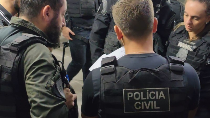 Observa-se um grupo de agentes da Pol&iacute;cia Civil em ambiente interno, vestidos com uniformes t&aacute;ticos e coletes de prote&ccedil;&atilde;o. Eles est&atilde;o reunidos de forma pr&oacute;xima, aparentemente analisando documentos ou discutindo detalhes de uma opera&ccedil;&atilde;o. A cena transmite organiza&ccedil;&atilde;o, foco e esp&iacute;rito de equipe, destacando o car&aacute;ter estrat&eacute;gico e colaborativo das atividades policiais.