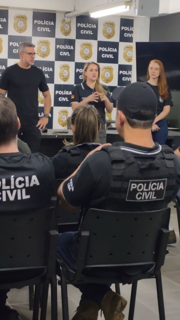 O cenário é uma sala de instrução ou treinamento. Vários policiais, também com uniformes da corporação, estão sentados em fileiras, atentos ao que três instrutores em pé apresentam diante de uma parede estampada com o logotipo da Polícia Civil. O ambiente sugere um briefing formal, voltado para capacitação ou alinhamento estratégico, reforçando a importância da disciplina, da comunicação clara e da preparação contínua dentro da instituição.
