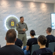 A imagem mostra o comandante-geral da Brigada Militar falando para um grupo de pessoas em um ambiente fechado. Atr&aacute;s dele h&aacute; uma parede branca, onde est&aacute; exposto o logo da Brigada Militar