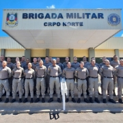 A fotografia colorida mostra um grupo de policiais militares em frente a uma sede da corpora&ccedil;&atilde;o. Detalhes da cena: O Grupo: Cerca de 25 policiais (homens e mulheres) est&atilde;o posicionados em forma&ccedil;&atilde;o para uma foto oficial. Eles est&atilde;o organizados em tr&ecirc;s fileiras horizontais, em degraus, todos em p&eacute;, com as m&atilde;os para tr&aacute;s e express&atilde;o s&eacute;ria. O Uniforme: Todos vestem o uniforme operacional da Brigada Militar do Rio Grande do Sul, composto por camisas e cal&ccedil;as em tom cinza-bandeirante, cintos pretos e botas pretas. O Pr&eacute;dio: Ao fundo, o grupo est&aacute; posicionado sob a marquise de um pr&eacute;dio de cor clara. Na fachada da marquise, l&ecirc;-se em letras pretas e grandes: "BRIGADA MILITAR", e logo abaixo, em letras menores, "CRPO NORTE". O bras&atilde;o oficial da corpora&ccedil;&atilde;o est&aacute; vis&iacute;vel &agrave; esquerda e &agrave; direita do letreiro. Ambiente e Ilumina&ccedil;&atilde;o: A foto foi tirada sob luz solar intensa e c&eacute;u azul com poucas nuvens. As sombras est&atilde;o projetadas diretamente abaixo dos policiais, indicando que o sol est&aacute; em uma posi&ccedil;&atilde;o elevada. H&aacute; rampas de acessibilidade com corrim&atilde;os brancos nas laterais do grupo.