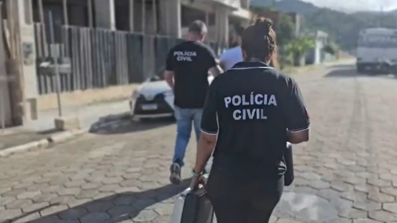 Dois policiais civis conduzem o preso por uma rua de piso tipo pave hexagonal