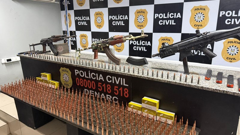 A imagem mostra uma apreens&atilde;o realizada pela Pol&iacute;cia Civil do Rio Grande do Sul, exibida em um balc&atilde;o diante de um painel institucional com o logotipo da corpora&ccedil;&atilde;o. Sobre a superf&iacute;cie est&atilde;o dispostos tr&ecirc;s rifles em posi&ccedil;&atilde;o vertical, diversos carregadores e uma grande quantidade de muni&ccedil;&otilde;es alinhadas em fileiras, al&eacute;m de caixas de proj&eacute;teis. A organiza&ccedil;&atilde;o cuidadosa dos itens sugere que se trata de uma apresenta&ccedil;&atilde;o oficial ou coletiva de imprensa, destacando o resultado de uma opera&ccedil;&atilde;o policial voltada ao combate ao crime e &agrave; circula&ccedil;&atilde;o ilegal de armas.