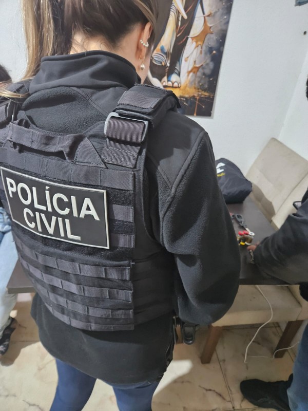 Na imagem, observa-se uma agente da Polícia Civil vestindo um colete tático preto com a inscrição “POLÍCIA CIVIL” em branco nas costas. Ela está em um ambiente interno, próximo a uma mesa onde se encontram diversos objetos pequenos, possivelmente materiais apreendidos ou evidências em análise. À direita, parcialmente visível, há outra pessoa também voltada para a mesa, sugerindo que ambos participam de uma atividade investigativa. Ao fundo, nota-se um cartaz na parede com uma figura estilizada, reforçando o caráter de cenário de operação ou investigação policial.