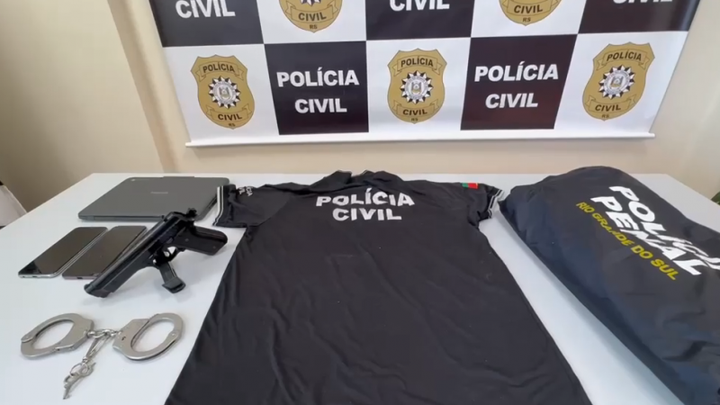 Na imagem, h&aacute; uma mesa posicionada diante de um painel com o logotipo e o texto &ldquo;POL&Iacute;CIA CIVIL RS&rdquo;, t&iacute;pico de coletivas ou apresenta&ccedil;&otilde;es oficiais. Sobre a mesa, est&atilde;o expostos diversos itens: uma camiseta preta com a inscri&ccedil;&atilde;o &ldquo;POL&Iacute;CIA CIVIL&rdquo;, um colete ou jaqueta escura com o texto &ldquo;POL&Iacute;CIA PENAL RIO GRANDE DO SUL&rdquo;, uma pistola, um par de algemas met&aacute;licas, dois celulares e um laptop. A disposi&ccedil;&atilde;o dos objetos sugere que se trata de materiais apreendidos ou equipamentos exibidos como parte de uma opera&ccedil;&atilde;o policial divulgada pela Pol&iacute;cia Civil do Rio Grande do Sul.