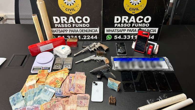 Uma mesa repleta de itens apreendidos pela Pol&iacute;cia Civil RS, especificamente da unidade DRACO Passo Fundo. Entre os objetos expostos est&atilde;o dois rev&oacute;lveres, muni&ccedil;&otilde;es, diversos celulares, balan&ccedil;as digitais, rolos de pl&aacute;stico filme, pacotes suspeitos, pen drives e uma quantia significativa de dinheiro em notas brasileiras. Ao fundo, dois pain&eacute;is pretos exibem o nome da institui&ccedil;&atilde;o e contatos, incluindo um n&uacute;mero de WhatsApp para den&uacute;ncias, refor&ccedil;ando o car&aacute;ter oficial da opera&ccedil;&atilde;o e a divulga&ccedil;&atilde;o p&uacute;blica dos resultados da apreens&atilde;o.