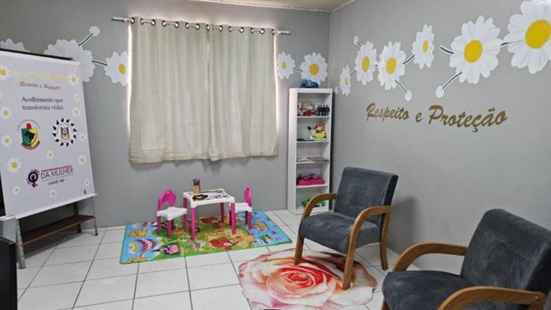 Esta &eacute; uma descri&ccedil;&atilde;o da imagem "Pol&iacute;cia Civil inaugura Sala das Margaridas em Quara&iacute;.jpeg" para fins de acessibilidade: A fotografia mostra o interior de uma sala de acolhimento humanizado, decorada em tons de cinza, branco e rosa, projetada para oferecer conforto e seguran&ccedil;a. Mobili&aacute;rio de Acolhimento: No centro e &agrave; direita da imagem, h&aacute; duas poltronas de estofado cinza escuro com bra&ccedil;os de madeira clara, posicionadas sobre um tapete circular que estampa uma grande rosa em tons de rosa e branco. Espa&ccedil;o Infantil: &Agrave; esquerda, sobre um tapete colorido com desenhos l&uacute;dicos, encontra-se um conjunto de mesa branca com pernas rosa e duas cadeiras pequenas (uma rosa e uma branca), destinado a crian&ccedil;as. Decora&ccedil;&atilde;o das Paredes: As paredes cinzas s&atilde;o decoradas com adesivos de grandes margaridas brancas com miolo amarelo, conectadas por tra&ccedil;os que simulam ramos. Na parede &agrave; direita, l&ecirc;-se a frase "Respeito e Prote&ccedil;&atilde;o" escrita em letras douradas com relevo. Ao fundo, uma janela &eacute; coberta por cortinas claras de tecido leve. Elementos de Apoio: Entre a janela e as poltronas, h&aacute; uma estante branca estreita com prateleiras contendo brinquedos e objetos decorativos. No canto esquerdo, um banner vertical branco exibe o nome "Sala das Margaridas", os logotipos da Pol&iacute;cia Civil e do Governo do Estado, al&eacute;m da frase "Acolhimento que transforma vidas" e informa&ccedil;&otilde;es da Procuradoria Especial da Mulher. A imagem transmite uma atmosfera de tranquilidade e cuidado, caracter&iacute;stica dos espa&ccedil;os voltados ao atendimento de mulheres e crian&ccedil;as em situa&ccedil;&atilde;o de vulnerabilidade.