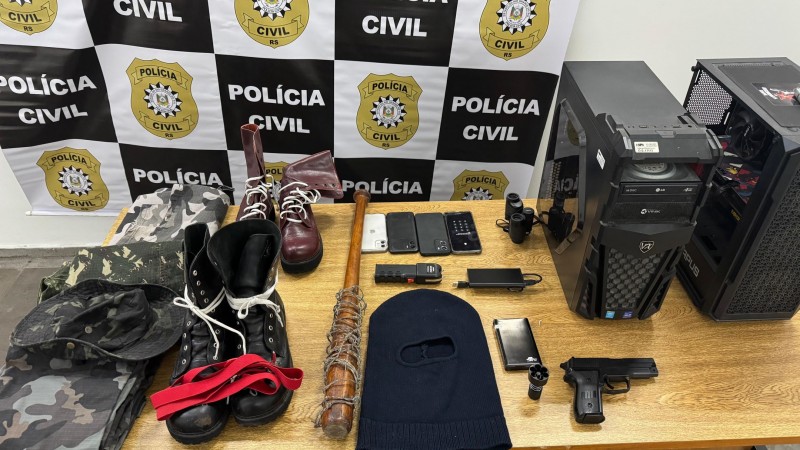 A imagem mostra uma mesa de madeira repleta de itens apreendidos pela Pol&iacute;cia Civil do Rio Grande do Sul, posicionada diante de um painel com o logotipo da corpora&ccedil;&atilde;o em quadrados pretos e brancos. Sobre a mesa est&atilde;o dispostos diversos objetos: roupas e chap&eacute;u camuflados, botas pretas e vermelhas, botas marrons, um bast&atilde;o de madeira envolto em arame, uma balaclava preta, uma pistola com carregador, celulares, um pendrive, uma lente de c&acirc;mera, al&eacute;m de dois gabinetes de computador. A cena transmite claramente o car&aacute;ter de uma exposi&ccedil;&atilde;o de evid&ecirc;ncias recolhidas em uma opera&ccedil;&atilde;o policial.