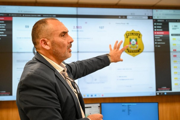 Em um ambiente tecnológico de monitoramento policial, um homem de terno gesticula diante de um grande painel digital repleto de gráficos, indicadores e o brasão da Policía Municipal com a bandeira do México ao centro. O cenário transmite a ideia de integração entre dados e segurança pública, sugerindo uma apresentação ou análise de operações policiais com apoio de ferramentas digitais.