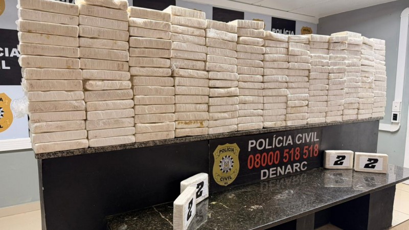 A imagem mostra uma grande quantidade de pacotes retangulares, todos embalados em pl&aacute;stico transparente, empilhados sobre um balc&atilde;o preto. Esse balc&atilde;o traz a inscri&ccedil;&atilde;o &ldquo;POL&Iacute;CIA CIVIL 08000 518 518 DENARC&rdquo; junto ao bras&atilde;o da corpora&ccedil;&atilde;o e as letras &ldquo;RS&rdquo;, indicando tratar-se da Pol&iacute;cia Civil do Rio Grande do Sul. Ao fundo, h&aacute; uma parede com o logotipo repetido da institui&ccedil;&atilde;o. O cen&aacute;rio sugere uma apreens&atilde;o significativa realizada pelo departamento antidrogas, com os pacotes expostos como evid&ecirc;ncia da opera&ccedil;&atilde;o.