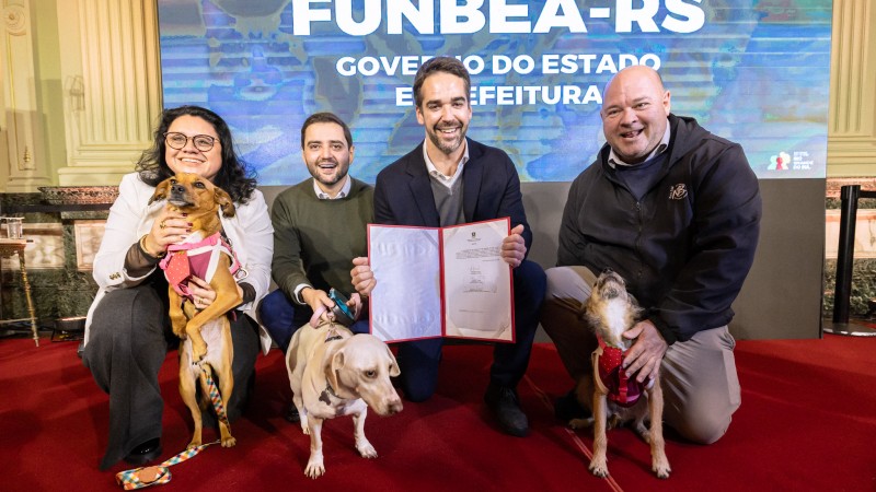 Grupo de quatro pessoas posando sobre um tapete vermelho em frente a um painel com o texto &ldquo;FUNBEA-RS&rdquo;, exibindo um documento aberto para a c&acirc;mera; tr&ecirc;s c&atilde;es aparecem em primeiro plano ao lado dos participantes.