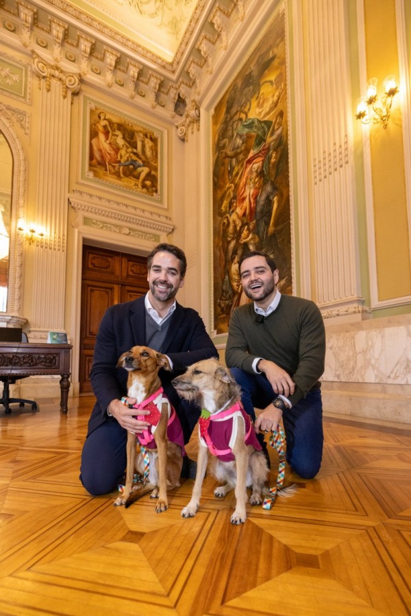 O governador e o vice-governador posam ajoelhadas ao lado de dois cães usando peitorais coloridos, em uma fotografia realizada no centro do salão.