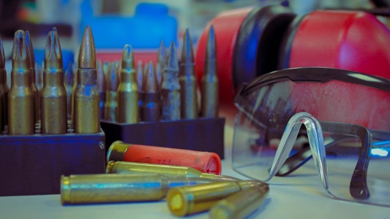 Foto mostra cartuchos de armas de fogo em estojos e espalhados sobre a bancada. Ao lado est&aacute; um &oacute;culos de prote&ccedil;&atilde;o.