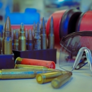 Foto mostra cartuchos de armas de fogo em estojos e espalhados sobre a bancada. Ao lado est&aacute; um &oacute;culos de prote&ccedil;&atilde;o.